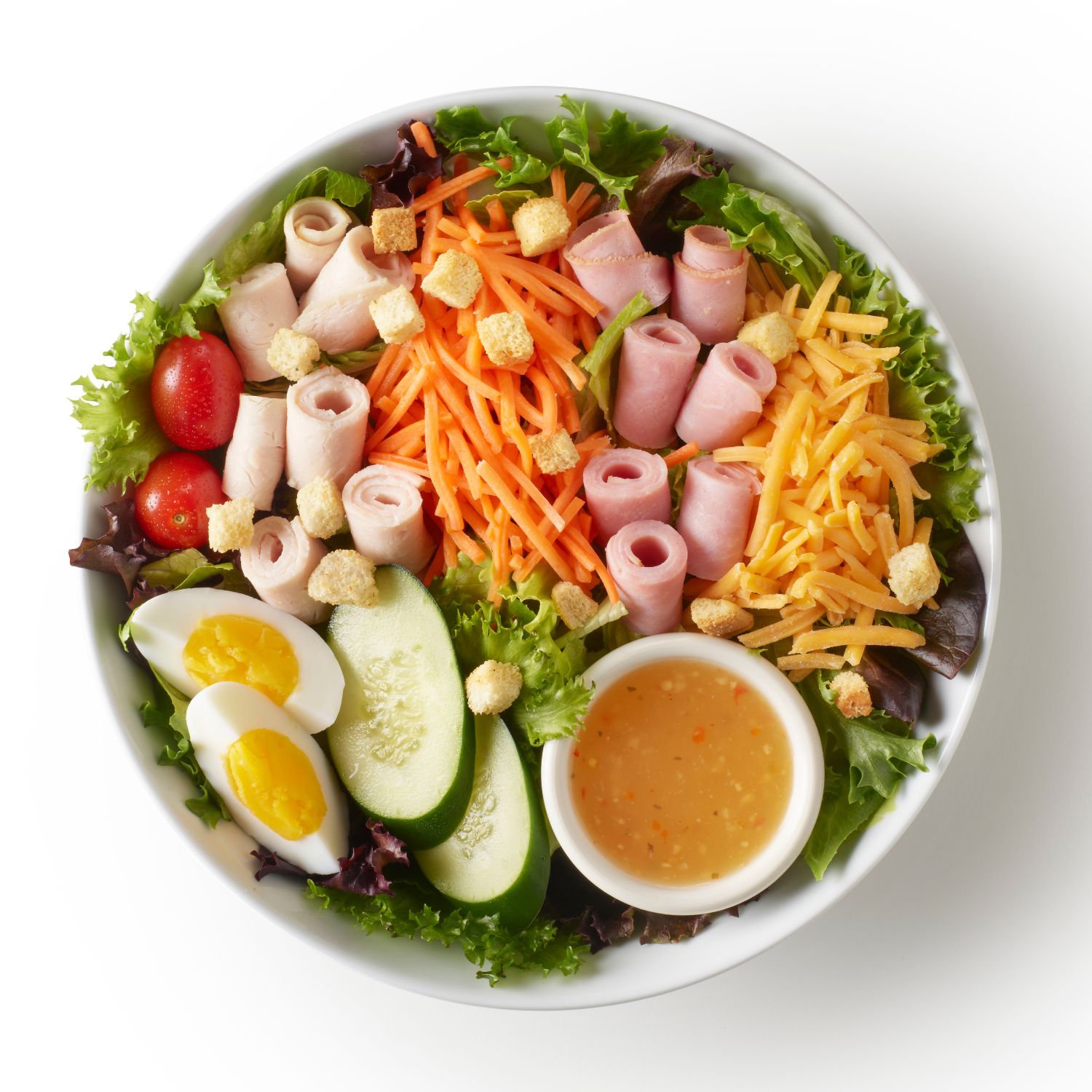 Top-down view of a chef salad with mixed greens, rolled turkey and ham, shredded cheese, carrots, eggs, tomatoes, cucumbers, croutons, and dressing.