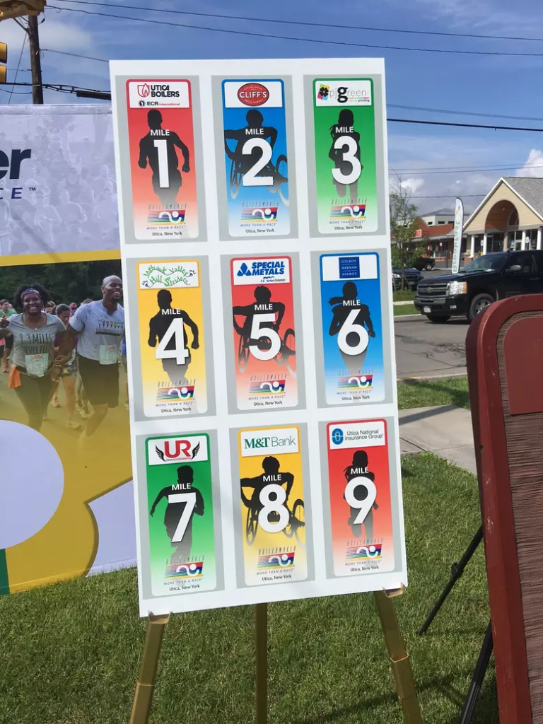 A Boilermaker Race sign with nine subsections for each mile in the overall 15 kilometer race. Each section red, blue, green, or yellow with a shadow silhouette of a racer. Featured is a sponsor above the racer's head and the Boilermaker logo in the bottom quarter of each image. The mentioned sponsors from 1-9 includes Utica Boilers, Cliff's Local Market, PJ Green, Mohawk Valley Hill Striders, Special Metals, Gilroy Kernan & Gilroy, Utica RoadRunners, M&T Bank, and Utica National Insurance Group.